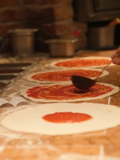 It all starts with the sauce. Here you can see our pizza assembly line where each hand-stretched base gets a generous layer of our rich, flavorful tomato sauce before the toppings go on.