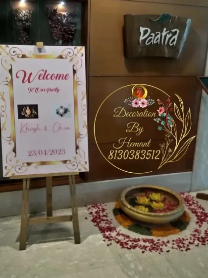 A classic welcome easel for a party, featuring a custom-designed welcome sign. The setup is complemented by a traditional brass urli with floating flowers and petals on the floor.