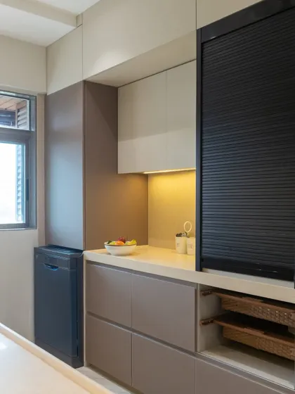 A view of the Vile Parle kitchen's utility area, showing the integrated dishwasher and a rolling shutter cabinet to neatly conceal small appliances. The open wicker basket shelves are perfect for easy access to everyday items.
