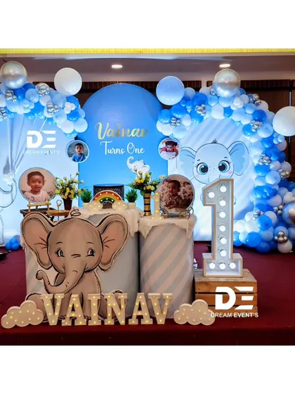 A complete stage setup for Vainav's baby elephant-themed first birthday. The design features a large arched backdrop, balloon garlands, and personalized elements like photo displays and name cutouts.