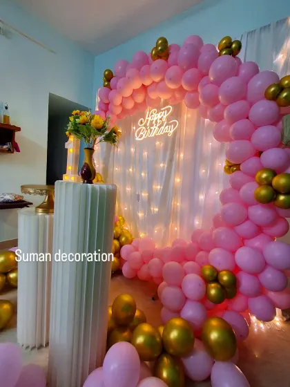 A pretty pink and gold birthday setup in a home. It features a balloon ring against a fairy light curtain, with plinths for the cake and flowers.