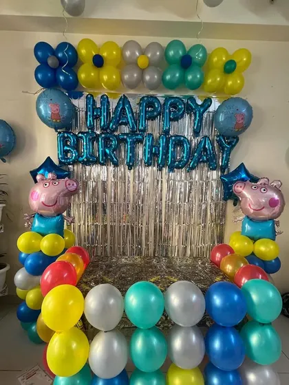 A colorful Peppa Pig themed decoration for a home celebration. The setup includes a silver shimmer curtain, 'Happy Birthday' foil letters, and Peppa Pig character balloons.