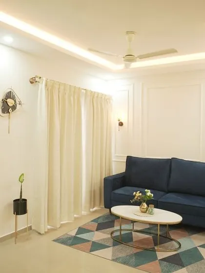 A clean and elegant living room with a blue sofa set against a white wall with decorative molding and floral wallpaper panels. The geometric rug adds a pop of color and pattern.