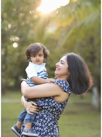 A candid photo of a mother holding her toddler in a park. The warm backlighting and her loving gaze create a warm and affectionate family portrait.