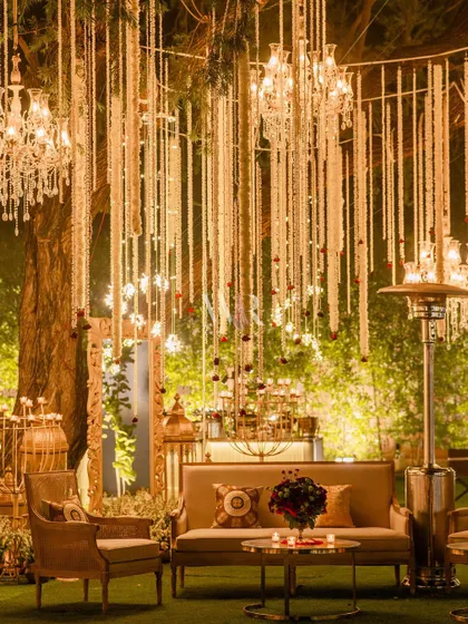 A wide shot of the sangeet lounge, showing the interplay of the natural tree, the hanging florals, and the warm lighting. The design seamlessly integrates the decor with the outdoor environment.