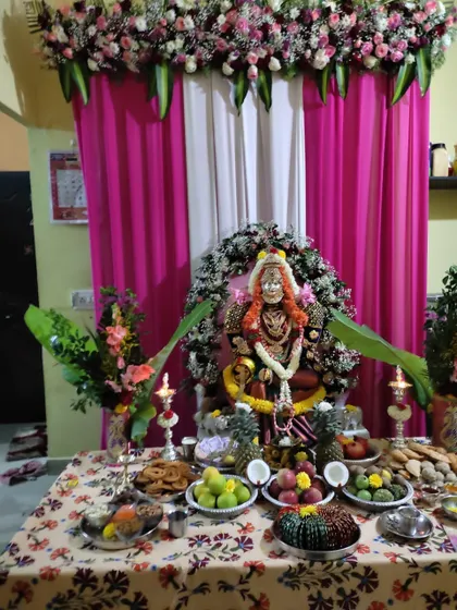 This is a home puja setup for a goddess deity. The decor includes a floral halo, banana leaf accents, and a backdrop of pink and white drapes.