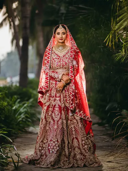 A classic, full-length portrait of the bride in her stunning red lehenga, set against a beautiful garden path. This image showcases the grandeur of her bridal attire.