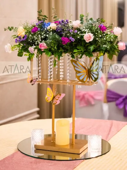 An elegant table centerpiece from a butterfly-themed event. A beautiful floral arrangement sits atop a golden stand adorned with butterfly cutouts and crystals, adding a touch of sophistication to the decor.