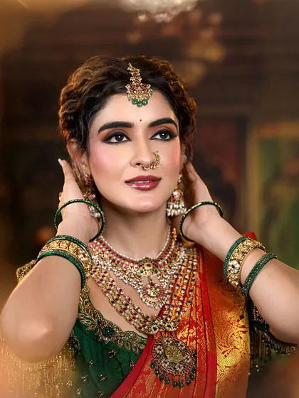 A regal and traditional bridal look featuring a rich red and green Kanjivaram saree. The makeup is defined by warm, shimmery eyes and a deep red lip, perfectly accessorized with temple jewelry.