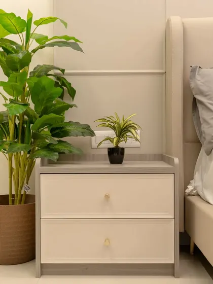 A detail shot of the bedside area, showing a two-tone nightstand and a large potted plant that adds a touch of nature to the room.