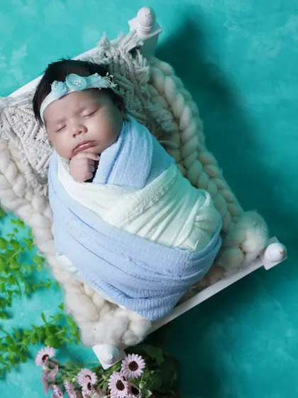 A top-down view of a newborn sleeping on a miniature bed. The turquoise background and surrounding flowers create a vibrant yet peaceful setting.