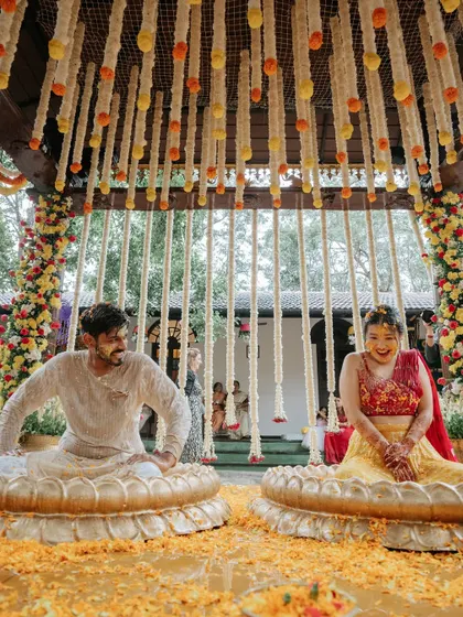 The couple seated in large lotus-shaped brass urlis, ready for the Haldi rituals. The mandap is decorated with hanging strings of tuberose and marigolds, creating a traditional and picturesque scene.