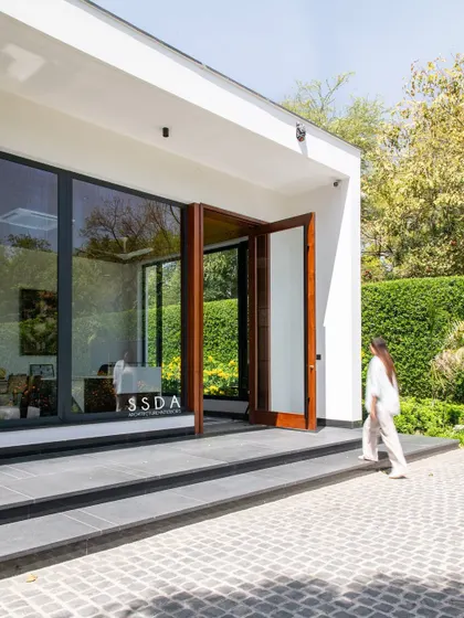 The entrance to The Green Axis office sets a tone of minimal elegance. The clean white facade and large wooden pivot doors are framed by a manicured hedge and stone steps, creating a welcoming threshold where architecture meets a controlled landscape.