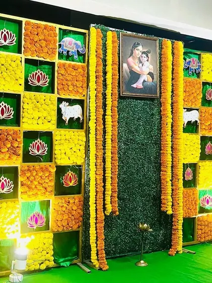 A vibrant marigold backdrop for a Seemantham ceremony. The checkerboard design includes auspicious symbols like elephants and cows, with a central portrait of a mother and child.