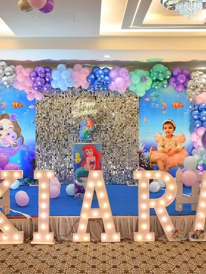 A complete Little Mermaid themed first birthday stage for Kiara. The setup includes an underwater backdrop, a silver shimmer wall, and large marquee letters.