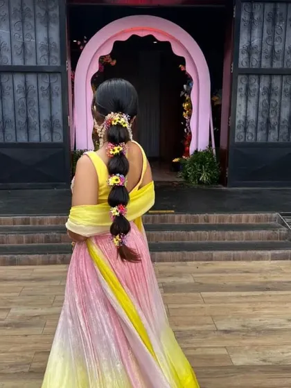 A full-length back view of a bride with a floral bubble braid, complementing her colorful ombre lehenga. It's a perfect hairstyle for a playful and festive Mehendi party.