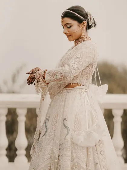 A bride in a stunning white lehenga, captured in a moment of quiet reflection on a balcony. The soft, overcast light adds to the serene mood.