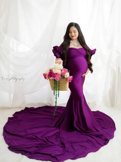 A regal and elegant portrait. The mom-to-be looks stunning in a deep purple gown, holding a basket of flowers against a clean white background.