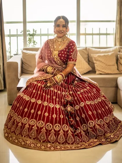 A full-length portrait of the bride seated, showcasing her magnificent red velvet lehenga. The makeup is designed to complement the richness of the fabric and the intricacy of the embroidery.