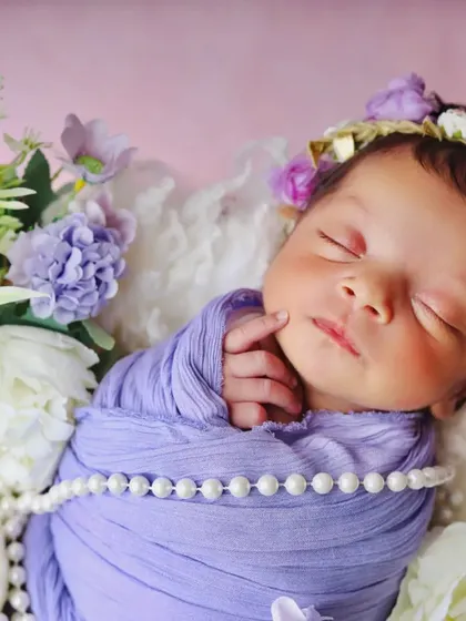 A close-up portrait of a newborn with a floral headband, peacefully asleep. The details of the pearl necklace and purple wrap add a touch of elegance.