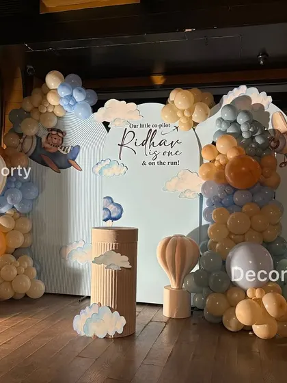 A charming 'Urso Aviador' or Aviator Teddy Bear themed first birthday setup. The backdrop features clouds, airplanes, and a large light-up number '1', surrounded by a soft blue and beige balloon garland.