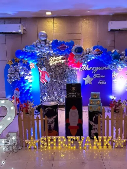 An out-of-this-world Space and Astronaut theme for a second birthday. This setup includes a silver shimmer wall, a crescent moon prop, astronaut cutouts, and a blue and silver balloon arch to create a cosmic adventure.