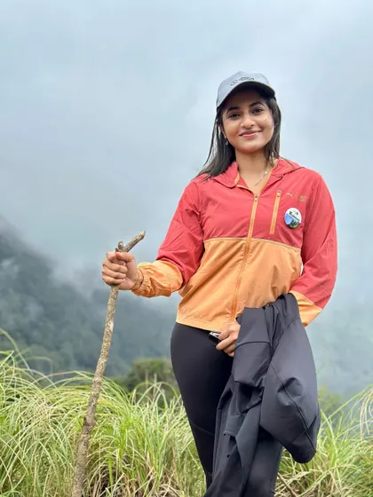 A happy trekker, geared up in her jacket and cap, holding a walking stick on the grassy slopes of Kurinjal.