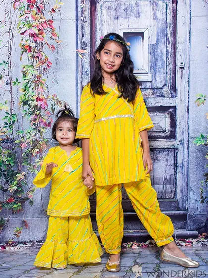 Two sisters in matching yellow traditional outfits hold hands for a lovely portrait. The coordinated clothing and their sweet connection make this a beautiful sibling photo.