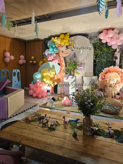 The main backdrop for the pastel jungle party features a friendly giraffe, a sleepy lion, and a soft color palette of pink, yellow, and mint green balloons.