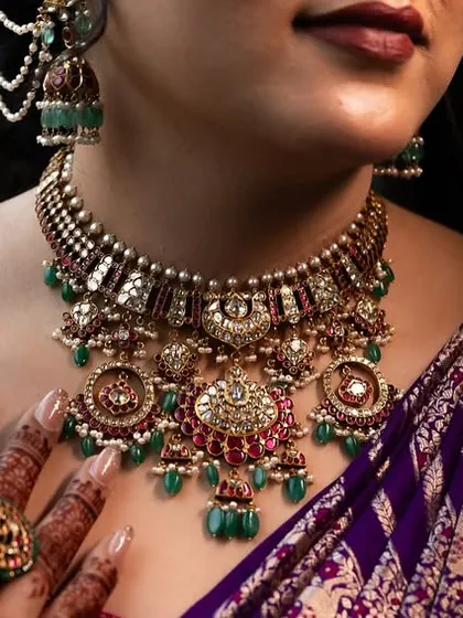 A close-up on the bride's neck, showcasing a magnificent, heavy bridal necklace with intricate carvings and mehendi-adorned hand.