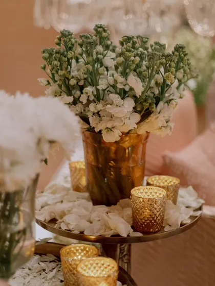 A close-up of the decor, showing simple arrangements of white flowers in textured gold vases, surrounded by candles. The petals scattered on the table add a touch of softness.