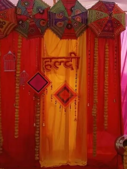 A traditional Haldi seating area with a backdrop of red and yellow drapes, decorated with umbrellas and marigold-filled pots.