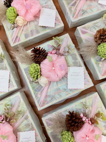 A top-down view of our Roka favors, ready for gifting. Each box is wrapped in a pastel-themed paper and adorned with a unique arrangement of fabric flowers, pinecones, and dried grasses.
