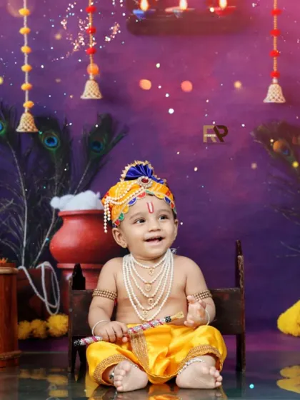 A smiling baby as Little Krishna, sitting on a small stool and holding his flute. The warm lighting and festive decorations create a happy and celebratory mood.