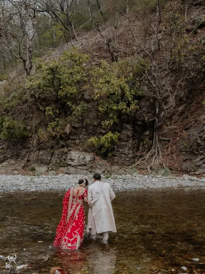 A beautiful wide shot of the couple wading through the river, showcasing the stunning Rishikesh landscape.