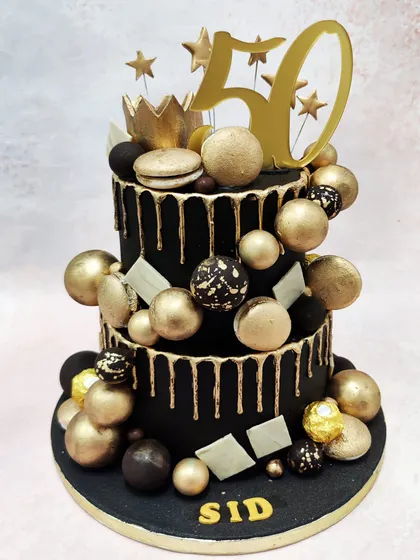 We mark a golden jubilee with this spectacular black and gold 50th birthday cake. Two tiers of black fondant with gold drips are lavishly decorated with a gold crown, stars, macarons, and a cascade of gold and black spheres for a truly grand celebration.