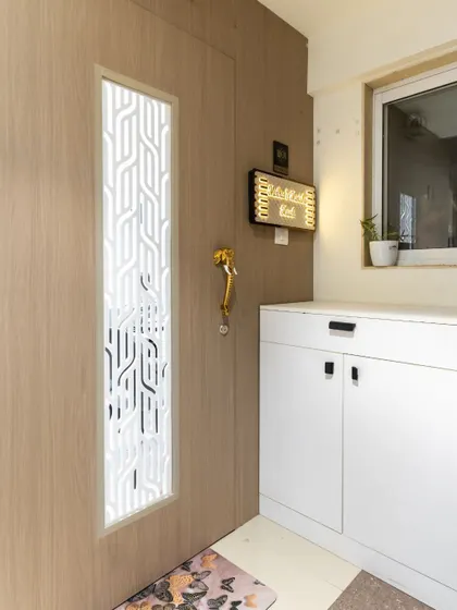This modern entryway features a light wood door with a geometric white jali panel. The design is complemented by a functional shoe cabinet and a beautiful nameplate.