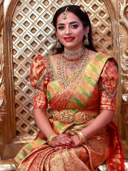 A truly royal bridal look. The bride is seated on a throne-like chair, wearing a multi-colored silk saree with puff sleeves. Her makeup is bold and elegant, with a red lip and diamond jewellery.