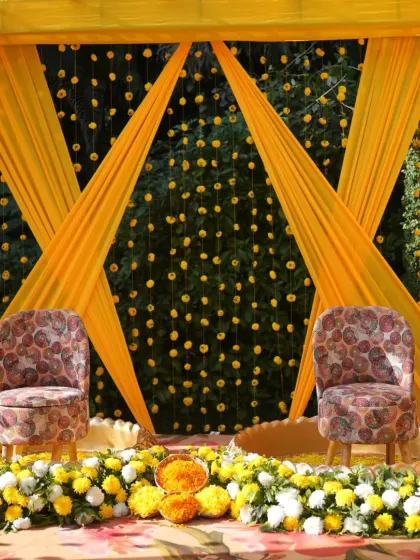 A traditional Haldi seating arrangement with a backdrop of crisscrossing yellow drapes and hanging marigold strings.