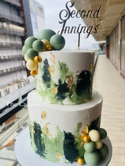 A modern and artistic two-tier cake for a 'Second Innings' or retirement party. The cake features abstract buttercream strokes in shades of green and black with gold leaf, decorated with chocolate spheres.