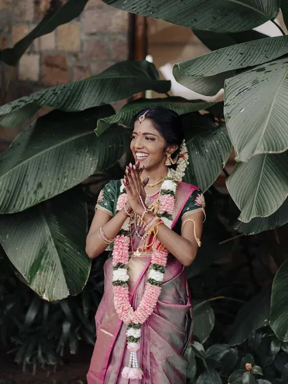 A joyful bride with her hands in a traditional gesture of greeting. The lush green background makes her pink saree and floral garland pop, capturing her vibrant personality.