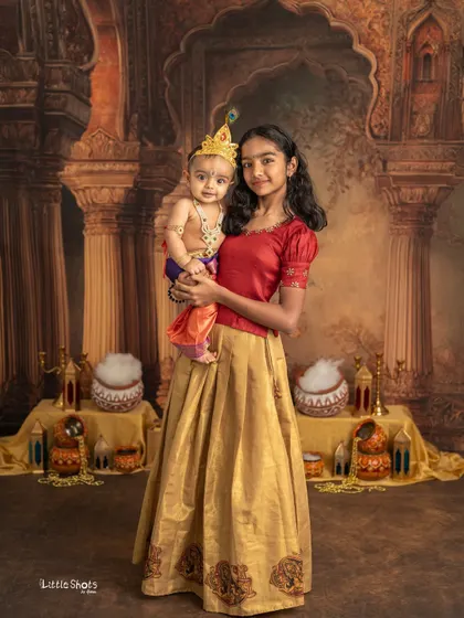 A full-length portrait of an older sister holding her baby brother, both dressed for a Janmashtami celebration. It's a beautiful picture of sibling love and tradition.