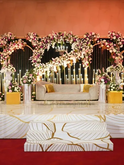 A grand and glamorous reception stage. The backdrop of candle-lit arches is framed by a cascade of pink and white flowers, with a stunning marble-patterned floor adding to the luxurious feel.