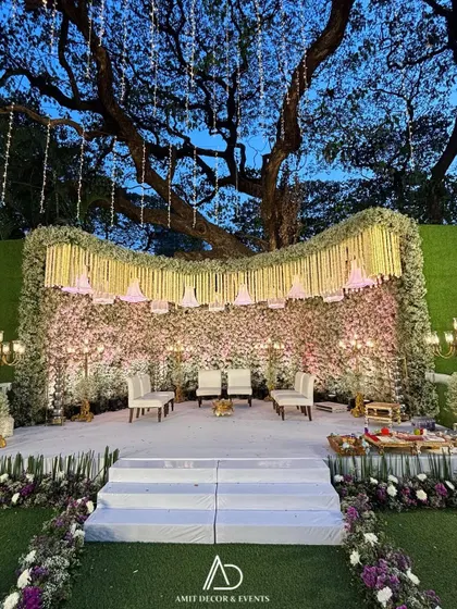 The main stage for an elegant evening event, featuring a wall of pink baby's breath, hanging lampshades, and a canopy of white floral strings under a majestic tree.