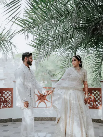 A beautiful portrait of the couple in coordinated white outfits, sharing a quiet moment under the shade of palm trees, showcasing a sophisticated pre-wedding look.