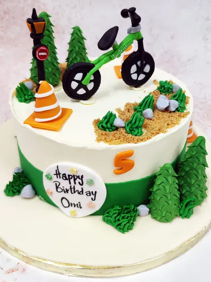 We set the scene for an outdoor adventure with this cycle theme cake. A green bicycle is parked on a dirt path surrounded by pine trees and traffic cones, creating a fun and detailed landscape for a young cyclist's birthday.
