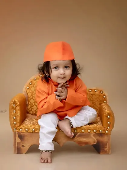 A minimalist portrait of a baby boy in a bright orange kurta. The solid background puts all the focus on his charming expression and curly hair.