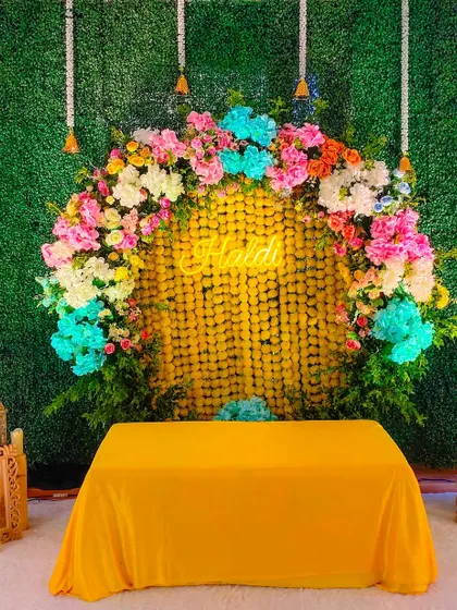 For a modern twist on Haldi decor, I created this backdrop using a green grass wall, a curtain of marigolds, and a vibrant multi-colored floral arch.