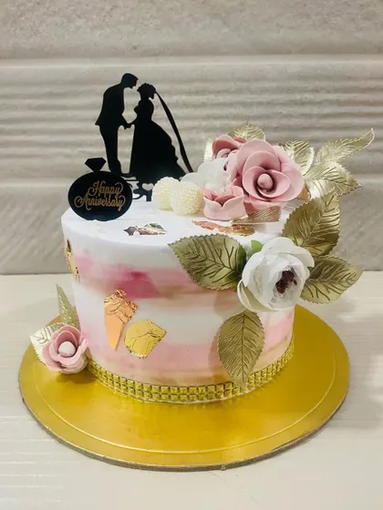 A romantic anniversary cake with a pink and white watercolor effect, adorned with golden leaves and a silhouette topper of a couple.