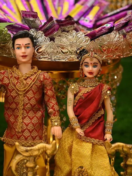 A pair of dolls representing the couple, perfect for an engagement, wedding, or seemantham ceremony. The background shows a plate of chocolates.